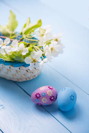 Two painted Easter eggs near basket with white spring flowering branch on light blue background.の写真素材