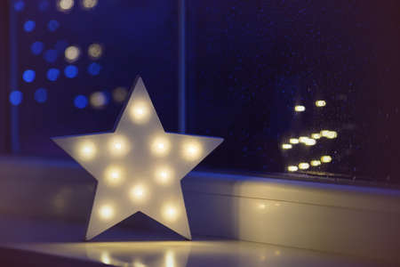 White LED star near window on garland bokeh background indoor in evening time. Festive spring illumination, holiday atmosphere.の写真素材