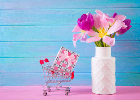 Small supermarket trolley with gift box and tender bouquet of beautiful pink tulips in vase on blue and pink wooden background. Concept of spring purchases.の写真素材