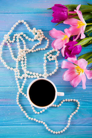Cup of coffee, white pearl necklace and tender bouquet of beautiful tulips on blue wooden background. Spring morning.の写真素材