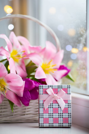 Gift box and tender bouquet of beautiful pink tulips in white basket near window with raindrops in...