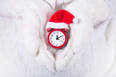 Closeup hands in white knitted mittens holding red alarm clock in small santa hat. New Year banner. Christmas card.の写真素材