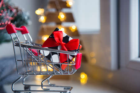 Pretty gift box in small supermarket trolley with warm garland lights on bokeh background. Concept of Christmas shopping.の写真素材