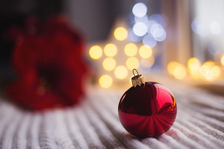 Red Christmas ball on white knitted fabric in daylight with warm garland bokeh on background. Festive magic lights. New Year holiday card.の写真素材
