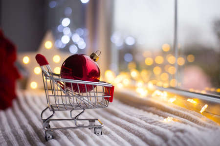 Red Christmas ball in toy supermarket trolley in daylight with warm garland bokeh on background. Festive magic lights. New Year shopping concept.の写真素材