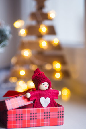 Little red Christmas doll with white heart in open gift box with warm garland bokeh on background. Festive romantic card.の写真素材