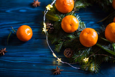 Wreath made from fresh mandarins with leaves and spruce branches with cones on navy rustic wooden table with garland and star anise. Christmas citrus decoration.の写真素材