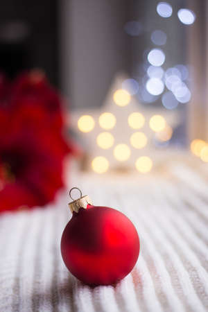 Red Christmas ball on white knitted fabric in daylight with warm garland bokeh on background. Festive magic lights. New Year holiday card.の写真素材