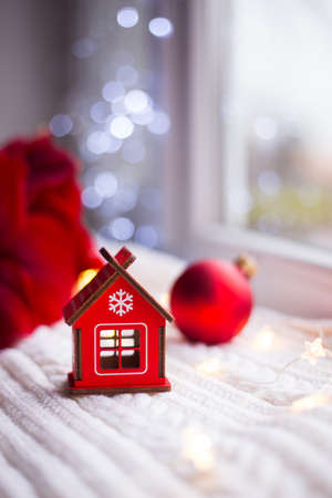 Toy red wooden house on white plaid surrounded with winter decor. Amaryllis, red Christmas balls, gift box and caramel canes near window in daylight. New Year holiday composition. Sweet home concept.の写真素材