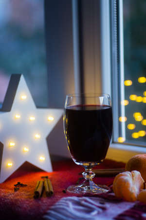 Mulled wine in glass surrounded by citrus, cinnamon sticks and cloves with LED white Christmas star and golden bokeh on background. Festive New Year composition near window.の写真素材