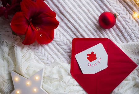 Christmas thankful greeting card in red envelope on white knitted plaid with Amaryllis flower, Christmas ball and glowing LED star.の写真素材