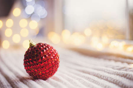 Red Christmas ball on white knitted fabric in daylight with warm garland bokeh on background. Festive magic lights. New Year holiday card with empty space.の写真素材