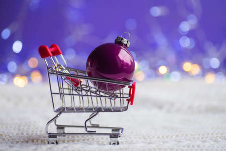 Crimson Christmas ball in small supermarket trolley on white knitted fabric with garland lights on purple bokeh background. New year banner with empty space. Concept of Xmas shopping.の写真素材