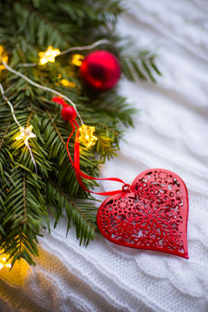Red heart with ornament near fir wreath decorated with Christmas balls and coiled with glowing garland with warm light on white knitted plaid. New Year home decoration. St. Valentine Day card.の写真素材