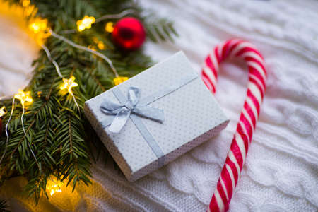 White gift box and caramel cane near fir wreath with red Christmas balls and coiled with glowing garland with warm light on white knitted plaid. New Year home decorationの写真素材