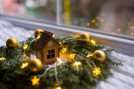 Toy wooden winter house near fir wreath decorated with golden Christmas balls and coiled with glowing garland with warm light on white knitted plaid. New Year home decorationの写真素材