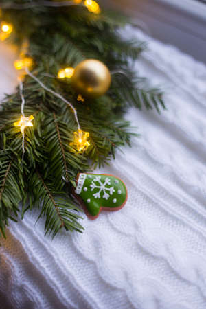 Toy little glove with snowflake near fir wreath decorated with golden Christmas balls and coiled with glowing garland with warm light on white knitted plaid. New Year home decorationの写真素材