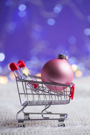 Pink Christmas ball in small supermarket trolley on white knitted fabric with garland lights on purple bokeh background. New year banner with empty space. Concept of Xmas shopping.の写真素材