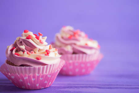 Closeup two cupcakes with creamy pink and white top decorated with little hearts on purple wooden background. St. Valentine day dessert.の写真素材
