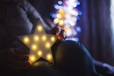 Female hands holding glowing white LED star with warm bokeh background indoor at home. Festive Christmas illumination, holiday atmosphere.の写真素材