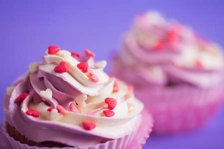Closeup two cupcakes with creamy pink and white top decorated with little hearts on purple background. St. Valentine day dessert.の写真素材
