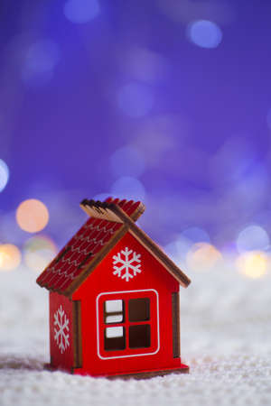 Red wooden house with snowflake on white knitted fabric on purple background with warm bokeh. New year card. Xmas decoration.の写真素材