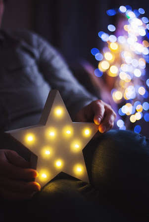 Female hands holding glowing white LED star with warm bokeh background indoor at home. Festive Christmas illumination, holiday atmosphere.の写真素材