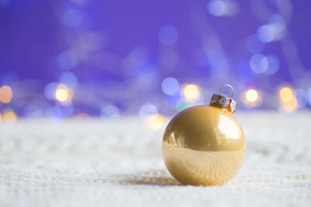 Golden Christmas ball on white knitted fabric on purple background with warm bokeh. New year card. Xmas decoration.の写真素材