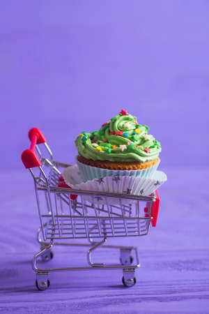 New Year cupcake with creamy green top with colorful stars and sprinkles in small supermarket trolley on purple background. Dessert in the form of Christmas treeの写真素材