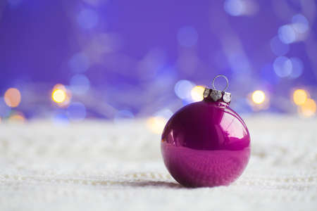Crimson Christmas ball on white knitted fabric on purple background with warm bokeh. New year card. Xmas decoration.の写真素材