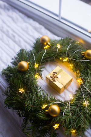 Golden gift box near fir wreath decorated with Christmas balls and coiled with glowing garland with warm light on white knitted plaid near window in daylight. New Year present.の写真素材