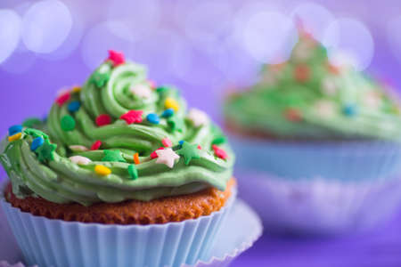 Closeup two New Year cupcakes with creamy green top with colorful stars and sprinkles on purple bokeh background. Dessert in the form of Christmas treeの写真素材