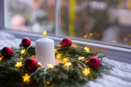Burning white candle in fir wreath decorated with red Christmas balls and coiled with glowing garland with warm light on white knitted plaid. New Year home decorationの写真素材