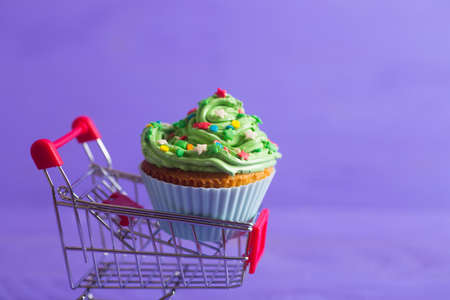 New Year cupcake with creamy green top with colorful stars and sprinkles in small supermarket trolley on purple background. Dessert in the form of Christmas treeの写真素材