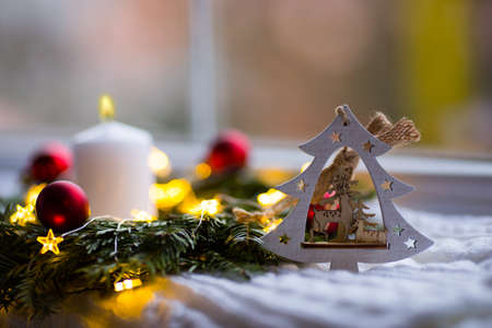 Decorative wooden Christmas tree with deer, burning white candle and fir wreath with red balls and warm garland lights near window. New Year home decor.の写真素材
