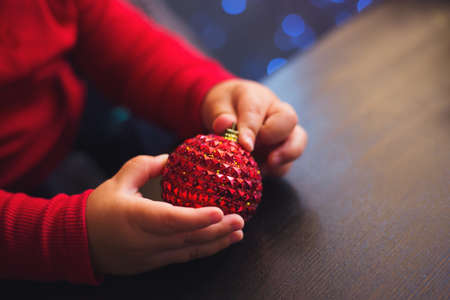 Children's hands holding red Christmas ball with garland bokeh background indoor at home. Toddler boy plays with New Year decoration.の写真素材