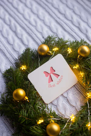 Greeting card with best wishes near fir wreath decorated with golden Christmas balls and coiled with glowing garland with warm light on white knitted plaid. New Year home decorationの写真素材
