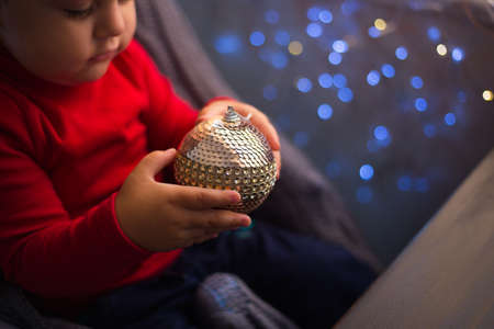 Children's hands holding shiny Christmas ball with garland bokeh background indoor at home. Toddler boy plays with New Year decoration.の写真素材