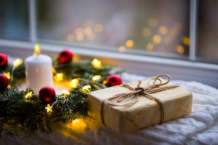 Wrapped gift box near fir wreath decorated with red Christmas balls, white burning candle and coiled with glowing garland with warm light near window in daylight. New Year presentの写真素材