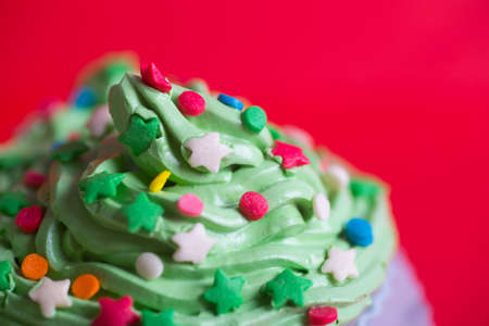 Closeup New Year cupcake creamy green top with colorful stars and sprinkles on red background. Dessert in the form of Christmas treeの写真素材