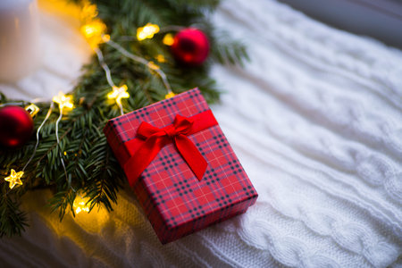 Closeup gift box near festive decoration with red Christmas balls and warm garland lights. New Year present.の写真素材