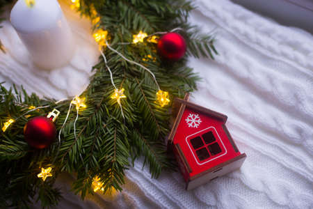 Toy wooden winter house near fir wreath decorated with red Christmas balls, white candle and coiled with glowing garland with warm light on white knitted plaid. New Year home decorationの写真素材