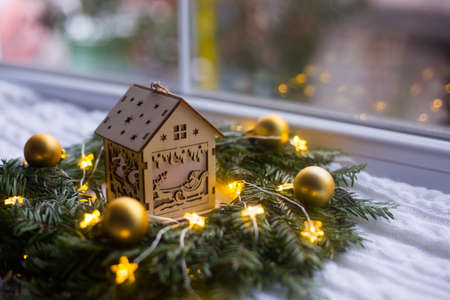 Toy wooden winter house near fir wreath decorated with golden Christmas balls and coiled with glowing garland with warm light on white knitted plaid. New Year home decorationの写真素材
