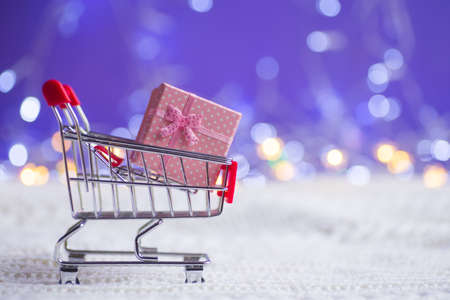 Pink gift box in small supermarket trolley on white knitted fabric on purple background with warm bokeh. New year banner with empty space. Concept of female or romantic giftの写真素材