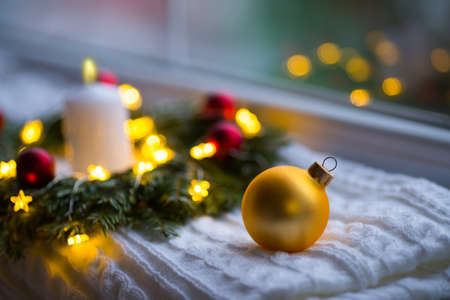 Golden Christmas ball near fir wreath decorated with white candle and warm garland lights near window. New Year cozy and festive atmosphere. Winter card with copy space.の写真素材