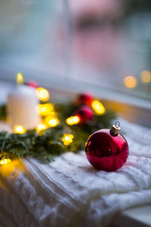 Red Christmas ball near fir wreath decorated with white candle and warm garland lights near window. New Year cozy and festive atmosphere. Winter card with copy space.の写真素材