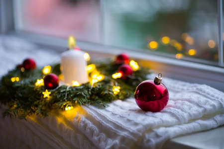 Red Christmas ball near fir wreath decorated with white candle and warm garland lights near window. New Year cozy and festive atmosphere. Winter card with copy space.の写真素材