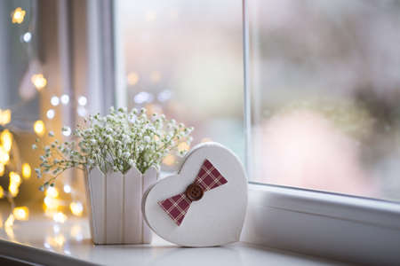 Heart shaped gift box and bouquet of beautiful white gypsophila near window in the daylight, bokeh lights on background. Tender romantic present.の写真素材
