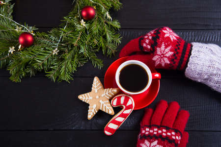 Hands in red winter gloves holding hot coffee cup with Christmas gingerbread on black wooden background. New Year homemade cookie with fir wreath and gift box.の写真素材