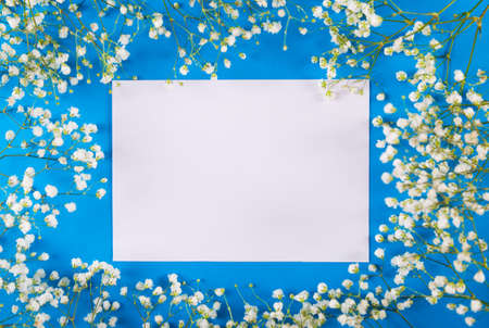 Empty sheet of paper surrounded with white gypsophila flowers on blue background, top view. Spring card with copy space for text.の写真素材